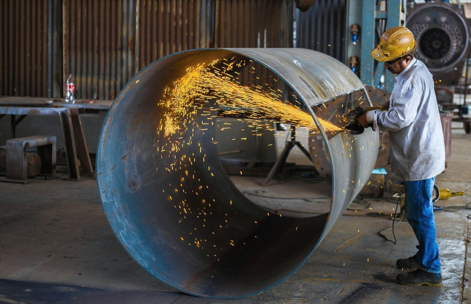 A worker in a hard hat grinding inside the mouth of a large rolled steel shell, sparks arcing across it