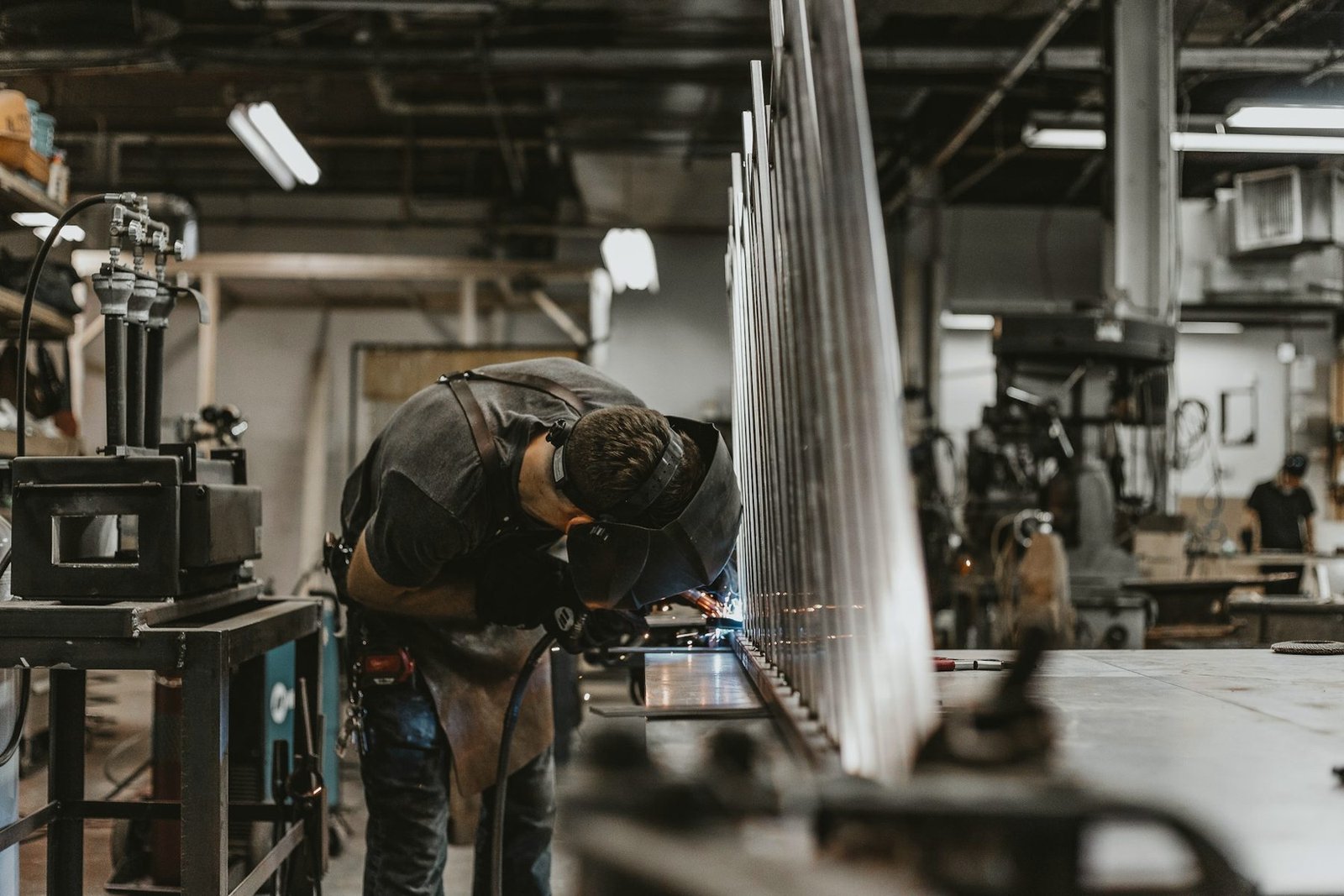 A fabricator bent over a work table in a busy metal shop, sheets of steel standing on edge beside him
