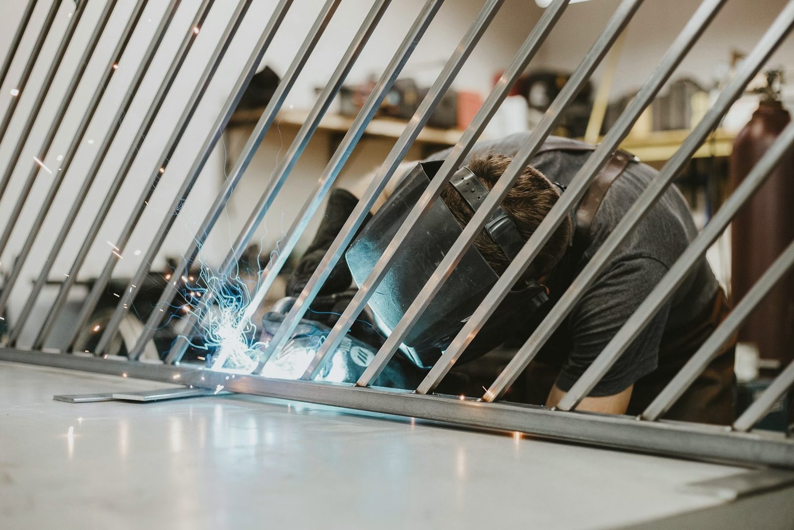A welder in a helmet seen through the angled pickets of a steel railing panel, welding a picket to the bottom rail